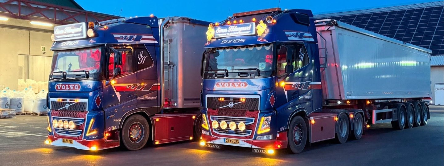 To blå Volvo-lastbiler fra Simon Holmsgaard Transport i skumringen med tændte lygter.