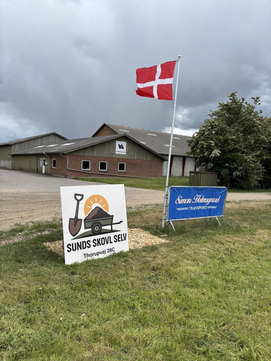 Skiltet til Sunds Skovl Selv på Thorupvej 28C med Simon Holmsgaard Transports blå banner og Dannebrog ved indkørslen til materialepladsen.